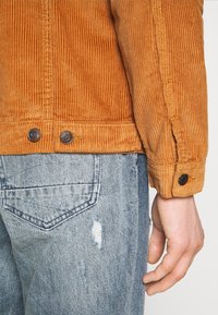 Veste en velours côtelé orange, présentant une texture côtelée, des boutons aux poignets, associée à un jean délavé bleu clair déchiré.