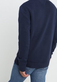 Granatowy dzianinowy sweter z fakturowanym wzorem, ściągaczami przy mankietach i dolnym brzegu, zestawiony z jasnoniebieskimi dżinsami.