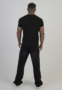 Musthave musta lühikeste varrukatega T-särk vastandlike äärtega, seljas tumedad, lõtvad teksad ja valged spordijalatsid, käed on paljad ja nähtavad tätoveeringud.