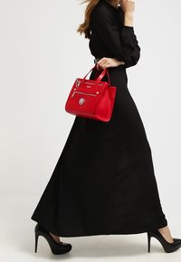 Borsa a mano in pelle rossa con tasca zip, hardware argentato e doppi manici, esposta accanto a un lungo vestito nero e tacchi alti.