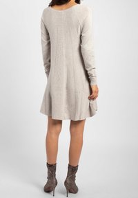 Femme portant une robe longue beige clair à manches longues, atteignant le genou, avec des panneaux en tricot verticaux, associée à des bottines à talons hauts scintillantes.