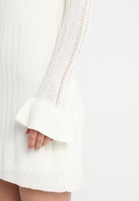 Abito bianco lavorato a maglia con texture a coste, maniche lunghe con tagli verticali e polsini svasati. Primo piano su una mano all'orlo.