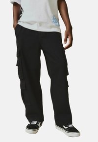 Pantaloni cargo neri con molte tasche laterali, vestibilità comoda e tessuto resistente. Indossati con una t-shirt grafica chiara e sneaker nere.