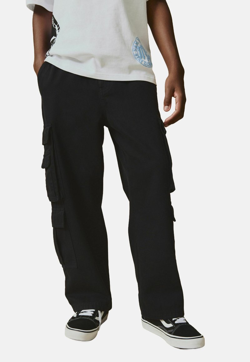 Pantalon cargo noir avec plusieurs poches latérales, en tissu robuste, associé à un t-shirt blanc ample et des baskets noires.