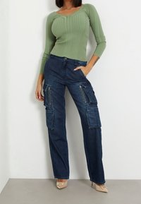 Gestreifter grüner Langarm-Top kombiniert mit Denim-Cargo-Hosen, die über mehrere Taschen und Reißverschlussdetails verfügen, ergänzt durch beigefarbene spitze Absatzschuhe.