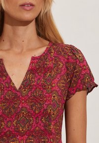 Kvinna som bär en kortärmad blus med intrikata geometriska mönster i rött, orange och brunt samt en v-ringning mot en enfärgad bakgrund.