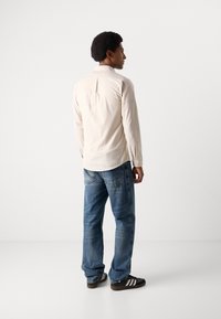 Ανοιχτό καφέ πουκάμισο με μακρύ μανίκι και γιακά με κουμπιά, με λεπτομέρεια πτυχής στην πλάτη, σε συνδυασμό με μπλε τζιν παντελόνι και μαύρα παπούτσια με λευκές ρίγες.