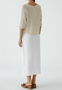 Top beige in maglia con maniche a ¾, abbinato a una lunga gonna bianca in lino. Sandali con tacco a blocco in legno marrone completano l'outfit.