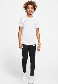 Maglietta bianca a maniche corte Nike abbinata a pantaloni sportivi neri Nike. Entrambi i capi presentano il logo Nike. Il modello indossa scarpe da ginnastica bianche.