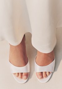 Sandali a ciabatta in raso bianco con una fascia larga sulla parte superiore, design con punta aperta, suola curva e texture liscia, su sfondo chiaro.