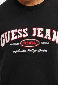 Sudadera negra con un llamativo texto en blanco y rojo. "GUESS JEAN" aparece de forma prominente junto con "1981" en rojo, todo dentro de un diseño ovalado. Textura suave.