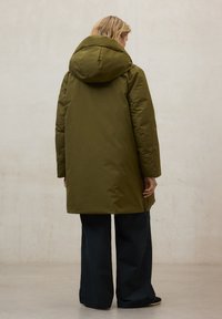 Chaqueta acolchada verde oliva con capucha grande, mangas largas y corte recto, combinada con pantalones anchos oscuros. Textura de tela suave.