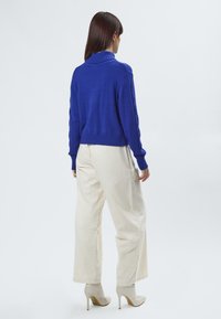 Blauer Strickpullover mit umschlagbarem Kragen, kombiniert mit cremefarbenen, weit geschnittenen Hosen und beigen High Heels; einfarbiger Hintergrund.