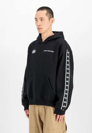 Hombre con una sudadera con capucha negra de Alpha Industries con logotipos en el pecho y las mangas y pantalones beige, de pie frente a un fondo blanco liso.