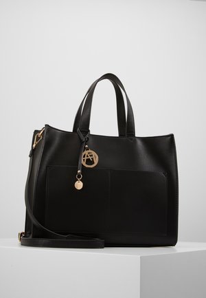 Sac fourre-tout en cuir noir avec deux poignées, poche avant, bandoulière amovible et pendentif logo circulaire doré suspendu à une poignée.