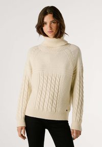 Krämfärgad stickad tröja med hög polokrage, prydd med kabelstickade mönster och ribbade manschetter och nederkant. Mjuk textur, avslappnad passform.