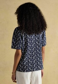 Tumedat sinine pluus lillemustriga, lühikeste varrukatega ja laineliste äärtega, valmistatud kergest kangast. Kantud heledate pükstega.