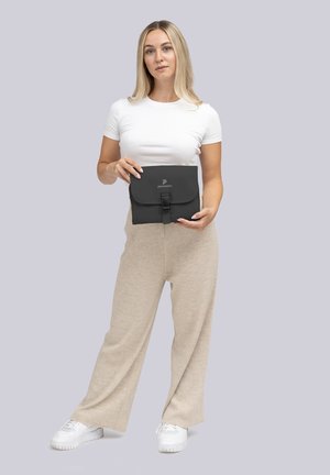 Bolso negro para tablet con cierre de solapa y detalle de hebilla, que presenta un logotipo. El modelo sostiene el bolso, vistiendo una camisa blanca y pantalones beige.