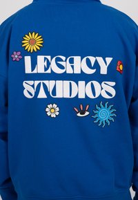 Mėlynas džemperis su užrašu "LEGACY STUDIOS" baltomis raidėmis, papuoštas spalvingomis gėlių ir grafikos aplinkybėmis įvairių formų ir dizainų.