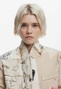 Collared shirt featuring a patchwork design in beige with various graphic patterns and prints. Soft fabric texture, front pocket detail.
