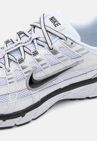 Adidas alb cu plasă și materiale sintetice, având accente negre, suprafață texturată și un sistem prominent de șireturi cu branding.