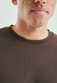 Brązowy bawełniany t-shirt z okrągłym dekoltem, z subtelnym logo „QS” w tym samym kolorze. Gładka tekstura, minimalistyczny design.