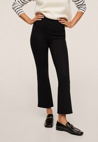 Pantalon noir à taille haute évasée, fabriqué dans un tissu extensible, avec des coutures latérales discrètes et une texture lisse. Assorti à des mocassins noirs.