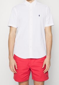 Vit kortärmad skjorta med knapp-up och en subtil randig mönster, med en liten blå logotyp. Kombinationen inkluderar ljusröda shorts.