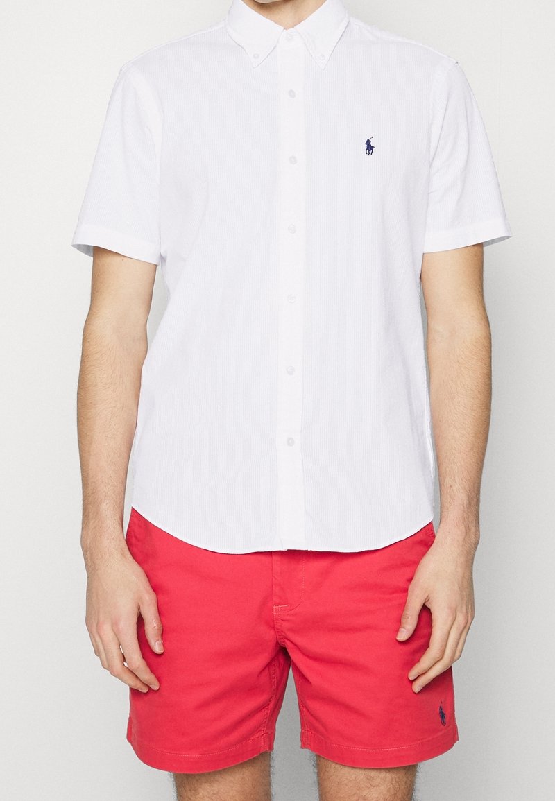 Vit kortärmad skjorta med knapp-up och en subtil randig mönster, med en liten blå logotyp. Kombinationen inkluderar ljusröda shorts.