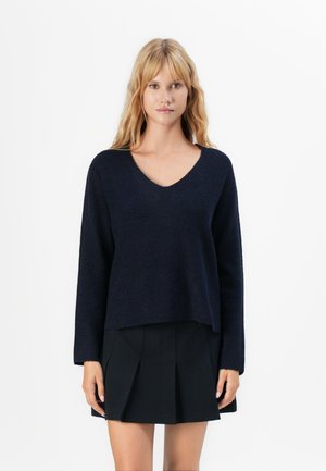 Woman with blonde hair wearing a navy blue V-neck sweater and black pleated mini skirt against a plain white background.