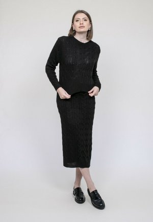 Femme debout portant un pull en tricot noir et une longue jupe assortie avec des mocassins noirs, sur un fond uni clair.