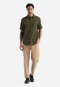 Olijfgroen kortemouwen shirt met een zak, gecombineerd met beige broeken en zwarte veterschoenen. Glad textuur en op maat gemaakte pasvorm.