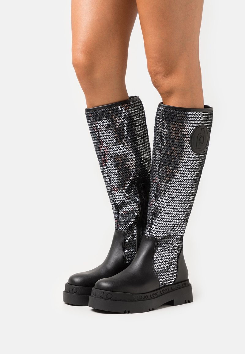 Bottines noires jusqu'aux genoux avec des panneaux de sequins brillants et des semelles en caoutchouc noires solides. Présente une fermeture éclair et un détail de logo en relief sur le côté.