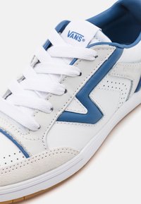 Baskets Vans blanches et bleues avec lacets blancs, détails perforés, bout en suède et semelle en caoutchouc visibles sur un fond blanc.