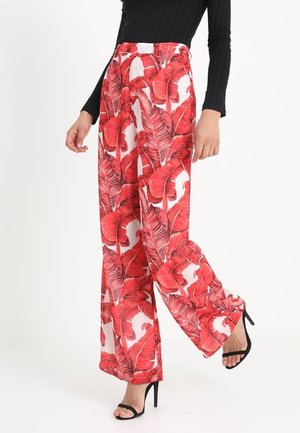 Mujer con pantalones de pierna ancha con estampado de hojas tropicales rojas intensas, combinados con una blusa negra de manga larga y sandalias negras de tacón alto.