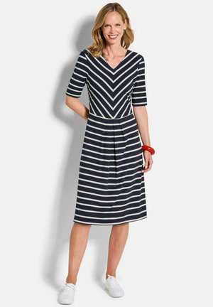 Femme portant une robe bleu marine et blanche à rayures, longueur genou, manches mi-longues, des baskets blanches et un bracelet rouge, debout et souriante.