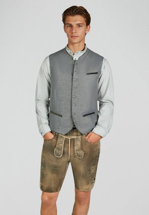 Junger Mann in traditioneller bayerischer Tracht mit grauer, geknöpfter Weste, hellem Hemd und bestickter brauner Lederhose, steht mit den Händen in den Taschen.