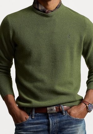 Mężczyzna w oliwkowo zielonym swetrze, kratcewej chuście, niebieskich dżinsach, brązowym skórzanym pasku i zegarku na nadgarstku, z rękami w kieszeniach, na jednolitym tle.