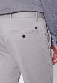 Primo piano della tasca posteriore di pantaloni grigi con bottone e piccolo logo a bandiera rossa, bianca e blu, abbinati a una camicia blu testurizzata.