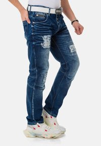 Blaue Jeans aus Denim mit beschädigten Stellen, schmaler Passform und Stickdetails. Kombiniert mit einem weißen Gürtel und Sneakers mit bunten Sohlen.