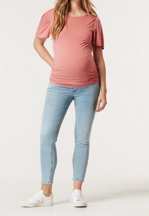 Osoba w ciąży ubrana w różowy top z krótkim rękawem, jasnoniebieskie dżinsy i białe trampki, stojąca z jedną ręką na biodrze.