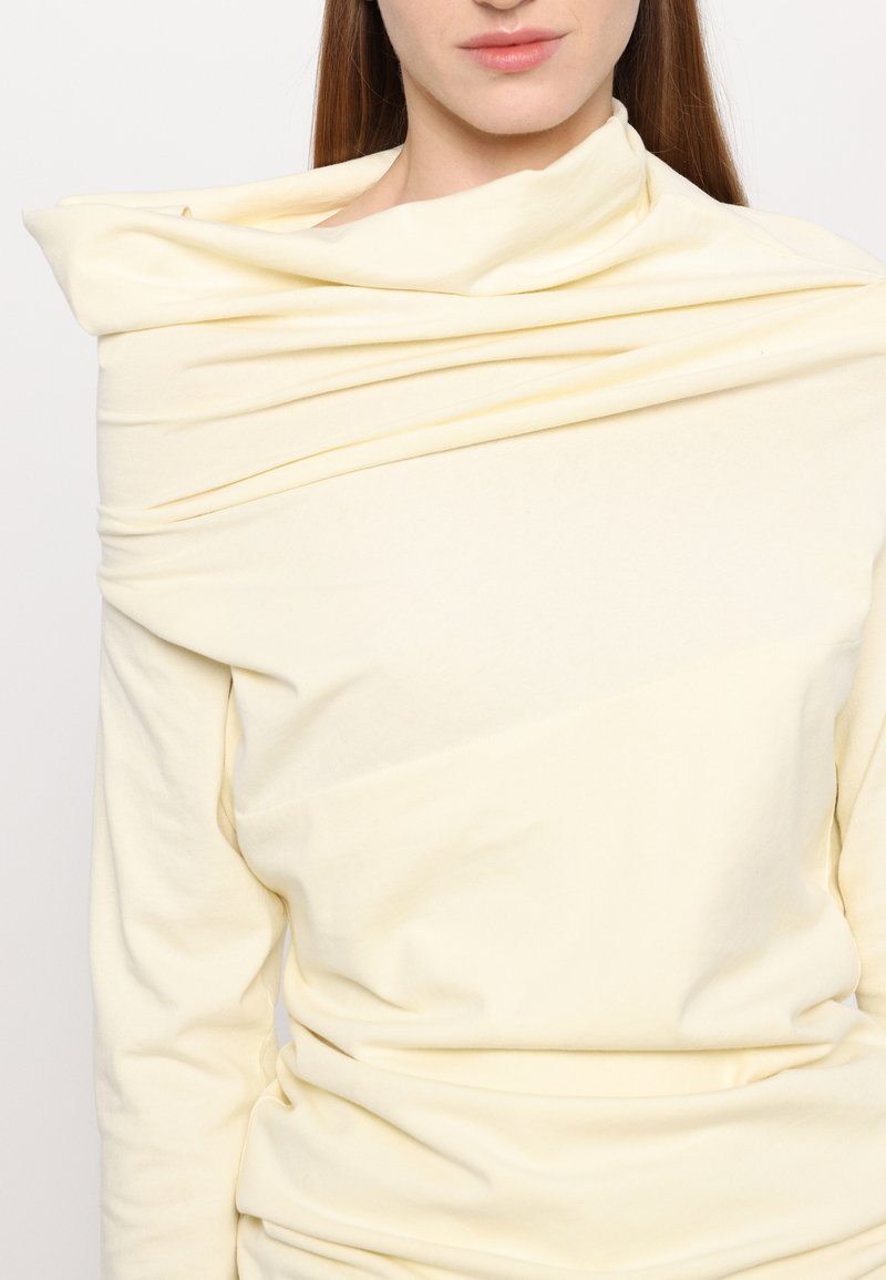 Krämfärgad långärmad topp med draperat tyg runt halslinningen, som har en mjuk, slät textur och minimalistisk design.