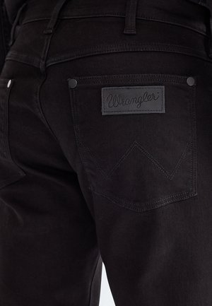 Primo piano della tasca posteriore di jeans denim neri con etichetta del marchio Wrangler e motivo "W" cucito.