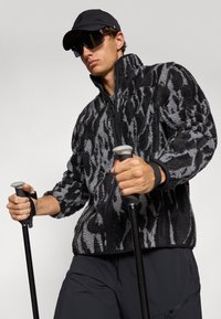 Veste en polaire noire et grise à motif, avec un col haut, fermeture éclair et surface texturée. Le mannequin tient des bâtons de randonnée, portant une casquette noire et des lunettes de soleil.