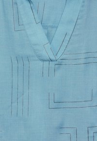 Camicia di tessuto blu con scollo a V, caratterizzata da sottili motivi lineari e accenti geometrici in toni più scuri. Texture morbida, materiale leggero.