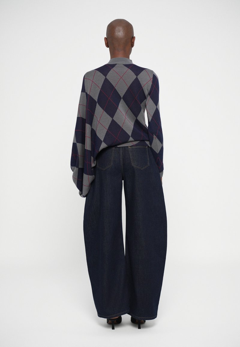 Maglione grigio a rombi con accenti blu navy e rossi abbinato a jeans in denim scuro a gamba larga. L'outfit presenta una vestibilità comoda e una texture morbida.