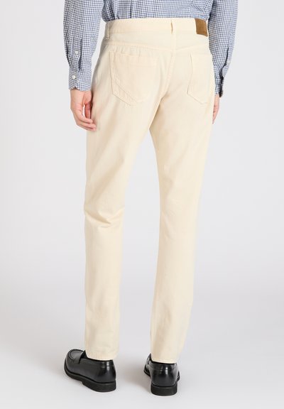 Pantalones de algodón beige claro con un corte recto, que cuentan con bolsillos traseros y un parche de cuero marrón en la cintura. Zapatos negros visibles.