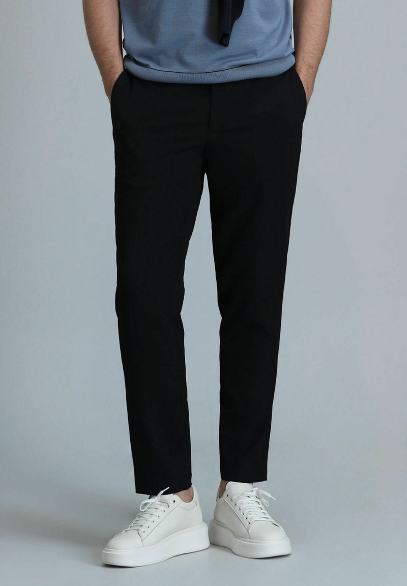 Pantalons noirs fuselés en tissu lisse, dotés de deux poches latérales et portés avec des baskets blanches sur un fond neutre.