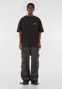 Sort bomulds t-shirt med broderi foran, parret med grå cargobukser med flere lommer og en afslappet pasform. Sorte sneakers fuldender looket.