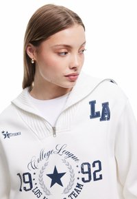 Suéter blanco con cuello alto y cremallera, con letras en color marino, estrellas y un estampado gráfico. Textura suave con detalle de cuello acanalado.